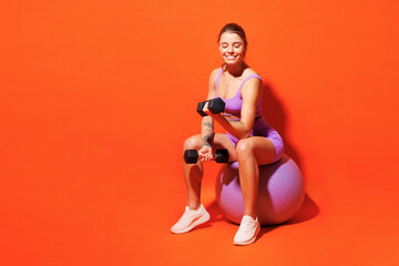 Full body happy young fitness trainer woman sportsman wear top shorts purple clothes train in home gym sit on fit ball hold dumbbells isolated on plain orange background Workout sport fit abs concept - 781902732
