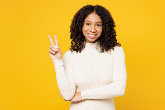 Little smiling happy cool kid teen girl of African American ethnicity wear white casual clothes showing victory sign isolated on plain yellow background studio portrait. Childhood lifestyle concept.
