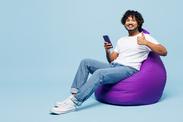 Full body young happy Indian man wear white t-shirt casual clothes sit in bag chair hold in hand use mobile cell phone show thumb up isolated on plain pastel light blue background. Lifestyle concept.