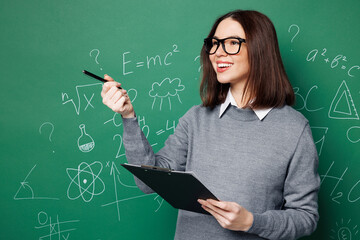 Young smart teacher woman wears grey casual shirt glasses clipboard with paper documents point pen aside isolated on green wall chalk blackboard background. Education in high school college concept.