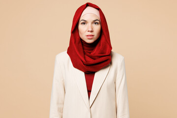 Young calm beautiful Arabian Asian Muslim woman wears red abaya hijab suit clothes looking camera isolated on plain beige wall background studio portrait. UAE middle eastern Islam religious concept.