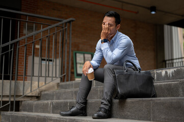 A stressed Asian businessman, appearing tired and unhappy, sits on the steps outside a building.