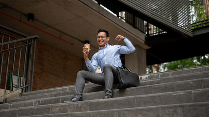A happy Asian businessman sits on steps, holding a coffee cup and cheering with a raised fist.