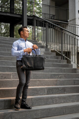 An exhausted Asian millennial businessman stands on the stairs, holding a coffee cup and a briefcase