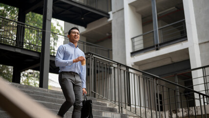 A confident businessman walks down stairs outside a building, holding a coffee cup and a briefcase. - 781921921