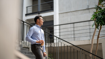 A confident Asian businessman walking down the stairs, holding a coffee cup and a briefcase.