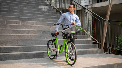 A focused Asian businessman pushing a green bicycle in the city, going to work with his bicycle.
