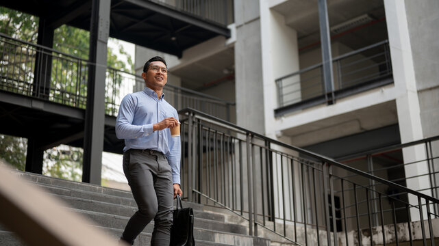 A confident businessman walks down stairs outside a building, holding a coffee cup and a briefcase.
