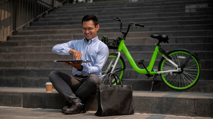 An Asian businessman sits on outdoor steps, opening his laptop, working remotely outdoors.