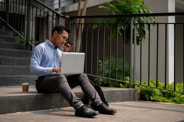 A businessman sits on steps, sending voice message via his smartphone while working on his laptop.