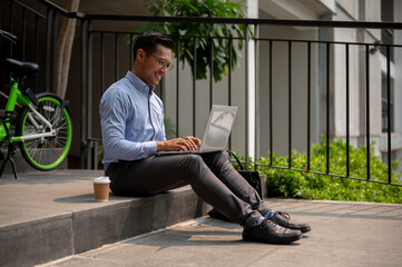 Confident Asian businessman sits on outdoor steps, smiling while using his laptop, working remotely.