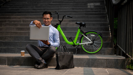 A happy Asian businessman sits on outdoor steps, smiling while using his laptop.