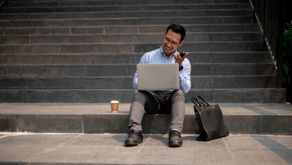 A businessman sits on steps, sending voice message via his smartphone while working on his laptop.