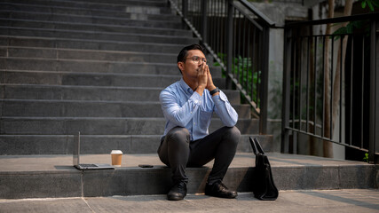An Asian businessman sits on outdoor steps, experiencing frustration and the fear of being fired.