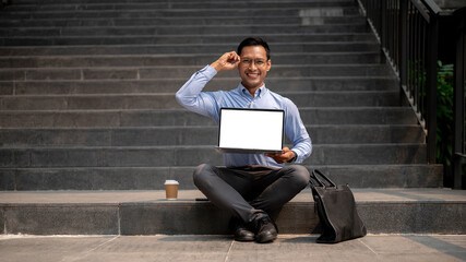 A confident Asian millennial businessman sits on outdoor steps, holding a laptop with a blank screen