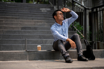An Asian businessman sits on steps, looking upward with his hand shielding his eyes from the sun.