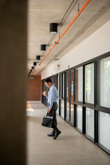 Determined Asian businessman walking along an indoor corridor, holding a coffee cup and a briefcase.