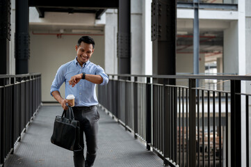 A confident Asian businessman is checking time on his wristwatch while walking along a corridor.