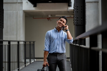 Confused Asian businessman talking on the phone with someone while standing on an outdoor corridor.