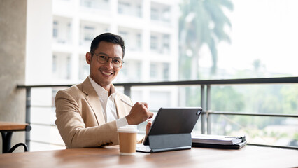 A confident, well-dressed Asian businessman sits at a wooden table with a tablet and documents.