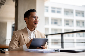 An optimistic Asian businessman smiles and looks away from the camera while holding a digital tablet
