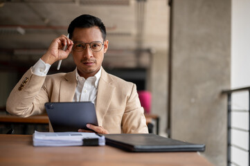 A confident Asian businessman touching his eyeglasses, looking at the camera, sitting at a table.