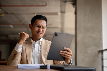 An enthusiastic Asian businessman celebrates with a clenched fist while looking at his tablet.