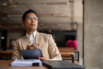 A shocked, amazed Asian millennial businessman holding a digital tablet and a pen, sitting at table.