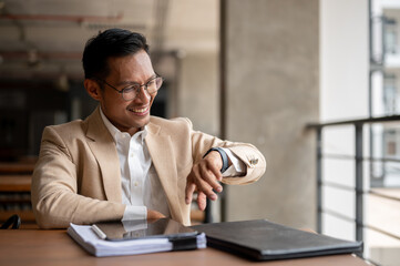A confident Asian businessman checking time on his wristwatch while sitting in a building corridor. - 781922993