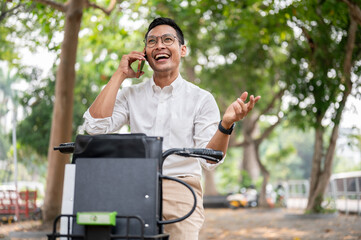 A happy Asian businessman is talking on the phone while he is on a bike, riding a bike to work. - 781923146