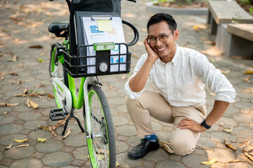 A happy, smiling Asian businessman sits next to his bicycle in a park, going to work in the morning.