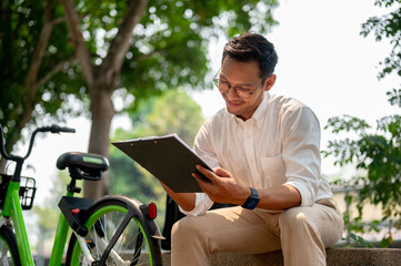 A confident, happy Asian millennial businessman reviewing his business document in a public park.