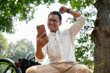 A cheerful Asian businessman sits outdoors, holding his phone with a triumphant expression. - 781923552