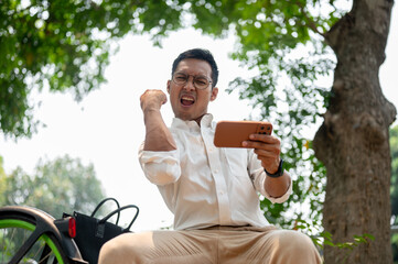 A cheerful Asian businessman sits outdoors, holding his phone with a triumphant expression.