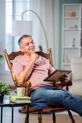 Indian asian mid age man looking at photo frame while sitting on rocking chair in living room