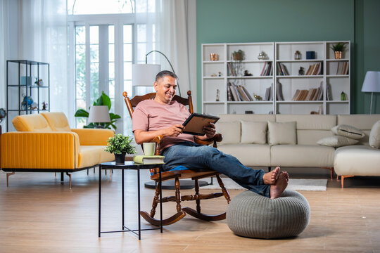 Indian asian mid age man looking at photo frame while sitting on rocking chair in living room - Powered by Adobe