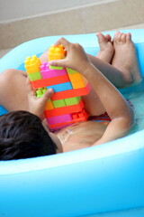 Cute little 3 year old boy is sitting in a sea blue bath tub and playing building blocks in the water. child is enjoying summer with water and making a building out of the colorful blocks. water time.