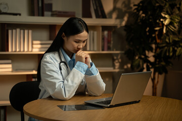 A young Asian doctor in a white coat is seated at a desk, a stethoscope around her neck,...