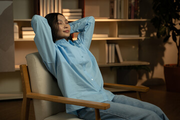 Fototapeta na wymiar A tranquil Asian woman sits comfortably in a cozy chair, her eyes closed as she stretches her arms in a relaxed gesture, surrounded by a warmly lit room with bookshelves, embodying peace and