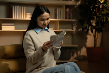 In a cozy home setting, an Asian woman concentrates on reading the instructions of her medication,...