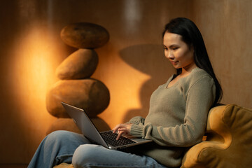 An Asian woman is seated in a chair, actively using a laptop computer. She is focused on the...