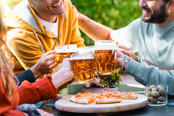 A group of friends cheer with beer mugs over a rustic table laden with pizza, enjoying a convivial...