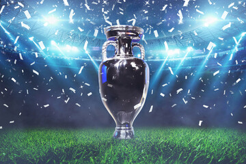 The champion cup stands on a green field at a football stadium with the light of spotlights and confetti. Football cup final, creative idea. Euro 2024 celebration - obrazy, fototapety, plakaty