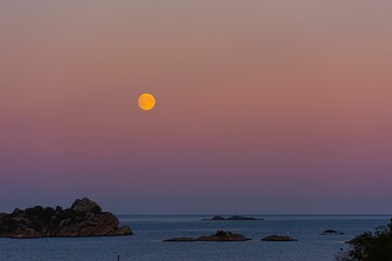 Full moon rising over the ocean in a pinkish and cloudless sky