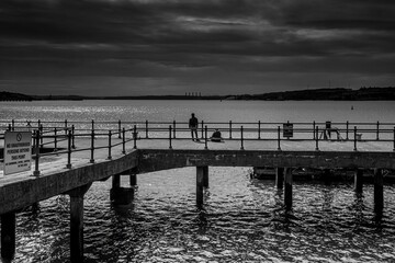 Fototapeta premium Scenic grayscale shot of people standing on a wooden pier and fishing in the tranquil sea