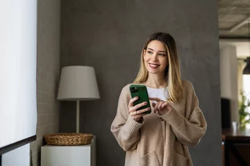 Tuinposter Young woman wearing sweater using smartphone at home, communication and social network concept, © NDABCREATIVITY