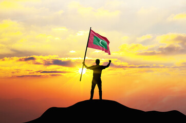 Maldives flag being waved by a man celebrating success at the top of a mountain against sunset or sunrise. Maldives flag for Independence Day.