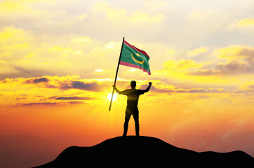 Mauritania flag being waved by a man celebrating success at the top of a mountain against sunset or sunrise. Mauritania flag for Independence Day.