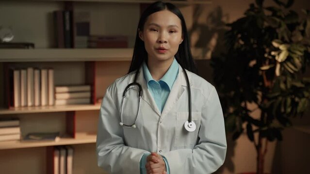 An Asian female doctor in a white coat with a stethoscope around her neck speaks directly into the camera, likely sharing important health information or providing a medical update. She stands