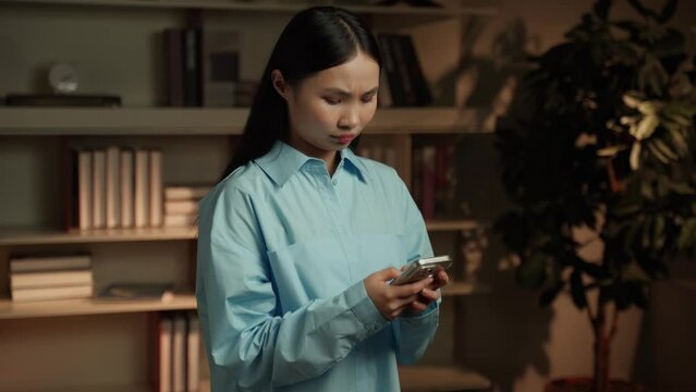 A young Asian woman is engrossed in her smartphone, browsing and interacting with the device, surrounded by bookshelves in a warmly lit home library as dusk settles outside.